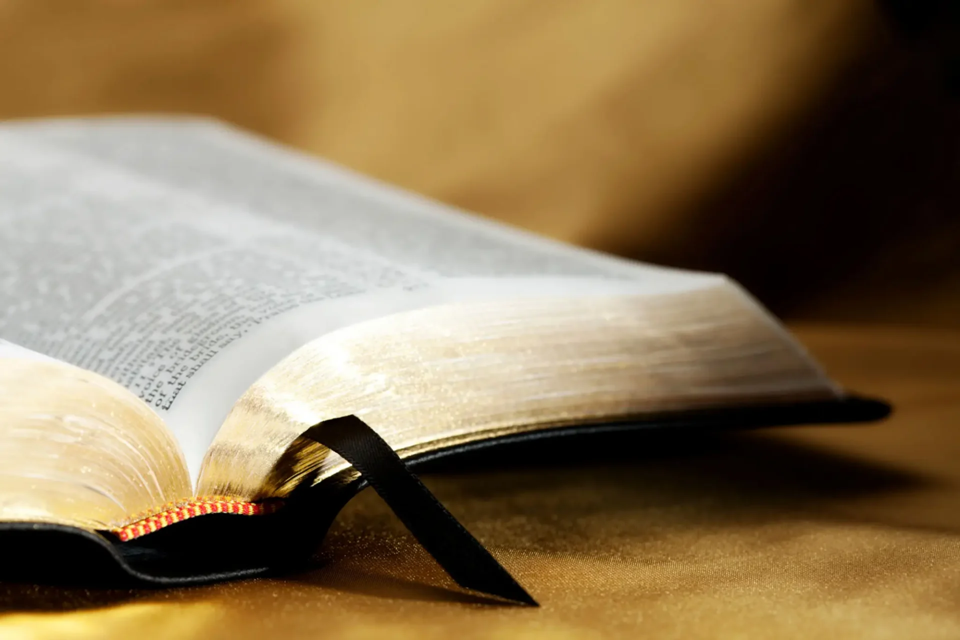 Open Bible with gilded pages and a black ribbon bookmark resting on a wooden surface.